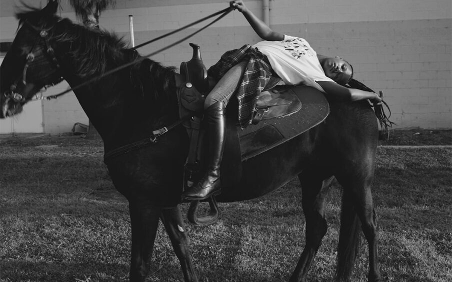 Morganne with Ebony. Melodie McDaniel. 2017. Photograph, archival pigment print. 33" x 44". Black and white photograph of a young Black individual posing on a tall black horse in a field of short grass in front of a light cement brick building with a palm tree to the left. The tall, thin rider is wearing black riding boots, jeans, a light colored t-shirt with undecipherable letters on it, and a plaid shirt tied at the waist. The rider is laid back in the saddle, back arched across the back of the horse, right arm gently extended holding the reins straight, and left arm outstretched, hand resting near the horse's tail along with the rider's ponytail of long braids. The tack has a simple design of thin leather straps and small silver buckles. The horse stands, left foot forward, head raised, mane blowing in the wind, with nose and mouth partially out of frame and slightly out of focus.