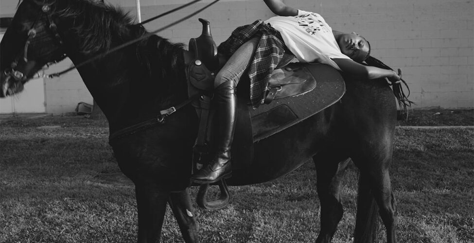 Morganne with Ebony. Melodie McDaniel. 2017. Photograph, archival pigment print. 33" x 44". Black and white photograph of a young Black individual posing on a tall black horse in a field of short grass in front of a light cement brick building with a palm tree to the left. The tall, thin rider is wearing black riding boots, jeans, a light colored t-shirt with undecipherable letters on it, and a plaid shirt tied at the waist. The rider is laid back in the saddle, back arched across the back of the horse, right arm gently extended holding the reins straight, and left arm outstretched, hand resting near the horse's tail along with the rider's ponytail of long braids. The tack has a simple design of thin leather straps and small silver buckles. The horse stands, left foot forward, head raised, mane blowing in the wind, with nose and mouth partially out of frame and slightly out of focus.