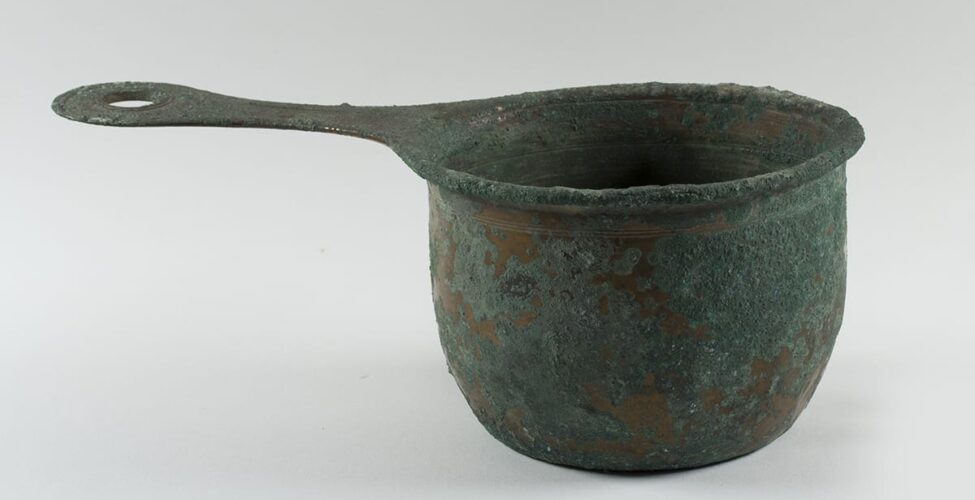 Saucepan, unknown Roman artist, 4 7/8 x 13 ¾ x 7 3/8 inches, bronze. A time worn pot with a rough, mottled, gray-green and bronze surface with a long, flat handle. The body of the flat-bottomed saucepan bows out slightly from its base and has a shallow lip that curves outward from the pot’s body. It has rough, crusty, gray-green corrosion on its surface with bits of smooth brownish bronze underneath showing through. A set of three very thin parallel grooves are incised around the pan’s top, just below the lip and more on the lip itself. More thin lines are seen on the interior of the saucepan. A flat handle protrudes from the left side of the pot. It narrows in the middle then widens into a rounded end with a circular cutout of its middle. More incised grooves form two circles around the cutout.