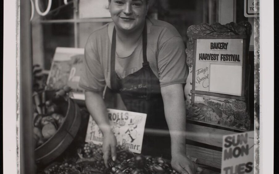 Kat, from the series North Portland Polaroids, Bobby Abrahamson, gelatin silver print, image: 13 5/16 x 10 1/16 inches; sheet: 14 x 10 15/16 inches. A portrait oriented, black and white photograph of an individual seen through a storefront window arranging produce and smiling directly at the viewer. Their dark hair is pulled back, and they have dark arched eyebrows, a dimple on their right cheek and a beauty spot on their left above their mouth. Their smile is warm and welcoming. They wear a short-sleeved t-shirt under a dark apron. Their hands reach down in front of them to arrange a large bowl of olives amid bunches of grapes, small boxes of cherries and other kinds of produce. The figure is flanked by small store signs. One elaborately framed one to the left reads “Bakery Harvest Festival”, to their right is an out of focus sign in the background and one is directly in front of the figure and advertises “Rolls” and “Thanksgiving Dinner”. At top left a neon sign is partially seen. It reads “…El Food” and “…O-Nuts”. Across the lower section of the photo, a horizontal streak of white is visible, possibly a light reflection on the store window. Behind the figure are elements of the shop: a paned glass window and doorway. A narrow uneven white line along the outer edge of the photo frames the scene.