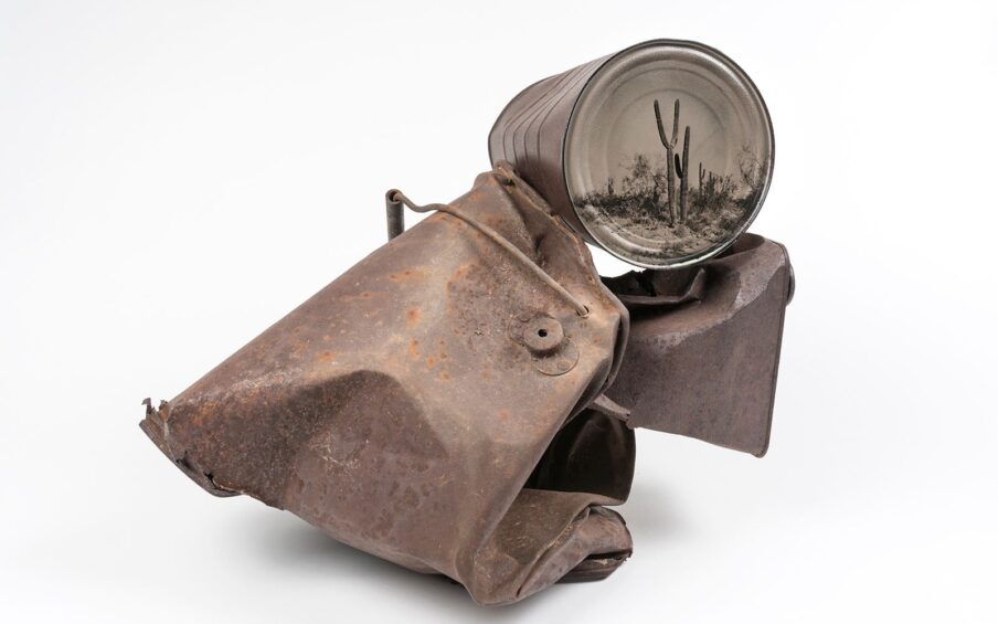 From the Conversations with History series, David Emmitt Adams. A sculpture composed of three main rusted metal objects, one containing a photo of a desert scene. A weathered, rusted tin can rests on its side on top of a crushed, dented metal container with a handle and a smaller piece of rusty trapezoid shaped metal. The end of the can has a black and white photo of two large cactus plants amid other vegetation in a desert setting. Below it sits the trapezoid shaped metal piece, rusted, the top of it crushed. Its original purpose is not identifiable. To the left is a larger metal piece, bigger than the other two pieces put together. It appears folded in half on itself, a sharp fold jutting towards the left with jagged edges. It has a handle with a grip bar and a small valve attached to its side with rivets. The metal pieces are rusty reddish brown and have a rough looking surface. Spots of orange rust speckle the body. The printed end of the can appears smoother and shows a bit of reflected light off its ridged surface.