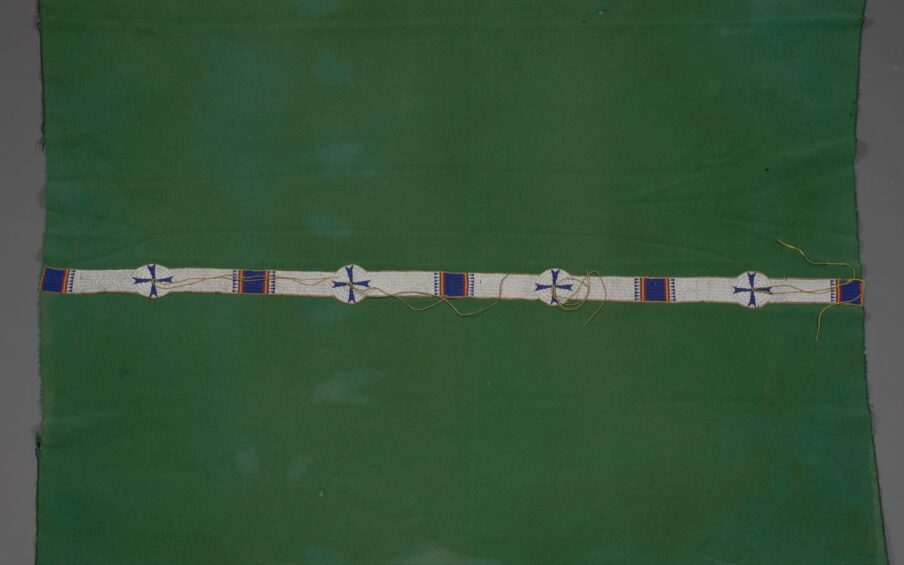 [Image description: Cheyenne artist, Blanket, 73 ¾ x 55 ½ inches, wool, glass beads. A rectangular, leaf green blanket with a narrow strip of beading running lengthwise along the middle. The blanket shows some discoloration where the fabric has faded and its edges are frayed where the stitched binding has come away. Stretching across the work, a narrow strip of white beading is punctuated by five blue beaded squares that are flanked on either side by slim yellow and red stripes and rows of blue beaded triangle shapes. Four round white beaded circles with blue crosses are interspersed between the squares. Pairs of long beige cords emanate from the circle centers, some lay stretched across the beading, some are coiled.]