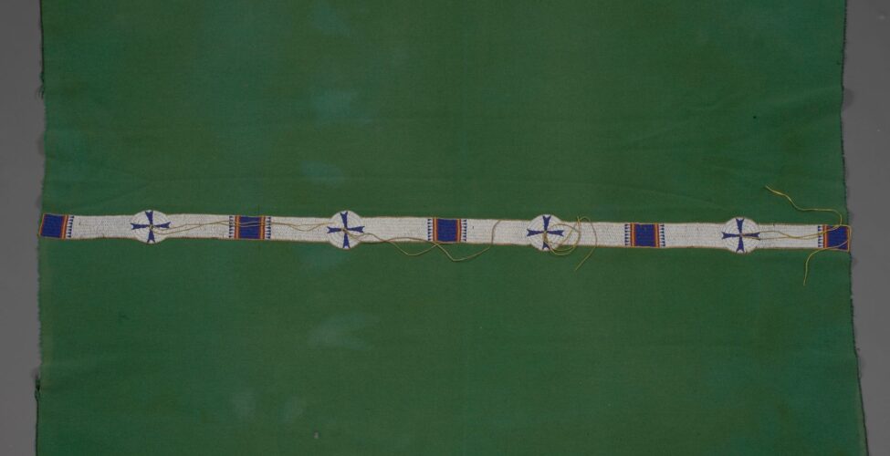 [Image description: Cheyenne artist, Blanket, 73 ¾ x 55 ½ inches, wool, glass beads. A rectangular, leaf green blanket with a narrow strip of beading running lengthwise along the middle. The blanket shows some discoloration where the fabric has faded and its edges are frayed where the stitched binding has come away. Stretching across the work, a narrow strip of white beading is punctuated by five blue beaded squares that are flanked on either side by slim yellow and red stripes and rows of blue beaded triangle shapes. Four round white beaded circles with blue crosses are interspersed between the squares. Pairs of long beige cords emanate from the circle centers, some lay stretched across the beading, some are coiled.]