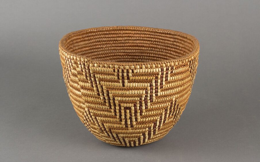 Image description: Basket, Cowlitz artist, cedar root, beargrass and dyed beargrass, 7 ¾ x 10 x 9 ¼ inches. 1. A round woven basket featuring a chevron pattern rendered in three tones of materials: light beige, medium brown, and dark brown. The basket has a wide mouth and gently narrows to a small base. It is tightly woven using the medium brown tone as a background on which the light beige and dark brown are used to create pattern. The dark brown and pale beige combine to repeat a double chevron pattern around the basket in an upper and lower row. At the center of each chevron at the top is a small, inverted triangle shape.