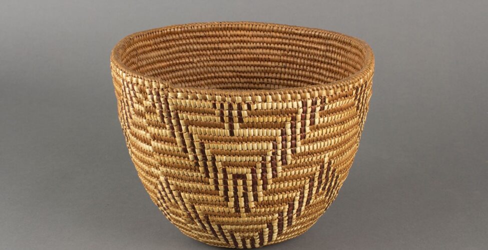 Image description: Basket, Cowlitz artist, cedar root, beargrass and dyed beargrass, 7 ¾ x 10 x 9 ¼ inches. 1. A round woven basket featuring a chevron pattern rendered in three tones of materials: light beige, medium brown, and dark brown. The basket has a wide mouth and gently narrows to a small base. It is tightly woven using the medium brown tone as a background on which the light beige and dark brown are used to create pattern. The dark brown and pale beige combine to repeat a double chevron pattern around the basket in an upper and lower row. At the center of each chevron at the top is a small, inverted triangle shape.