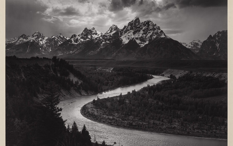 The Tetons and the Snake River, Grand Teton National Park, Wyoming, Ansel Adams, gelatin silver print. A landscape photograph showing the jagged peaks of the Grand Teton mountain range as a backdrop to the sharply curved Snake River in the foreground. The mountains begin a third of the way down from the top and stretch across the width of the photo. Snow-covered, jagged peaks overlap against a dramatic sky of clouds with sun breaking through. Below the mountains, low rolling hills meet the S curved river seen emerging from the vegetation at the center right. The river crosses diagonally to the left and switches back to the right at the bottom of the work. Steep scrub covered hills rise from the river edge at left. Low lying trees and vegetation fill in the right of the photo, while evergreen trees are seen at the bottom in the foreground.