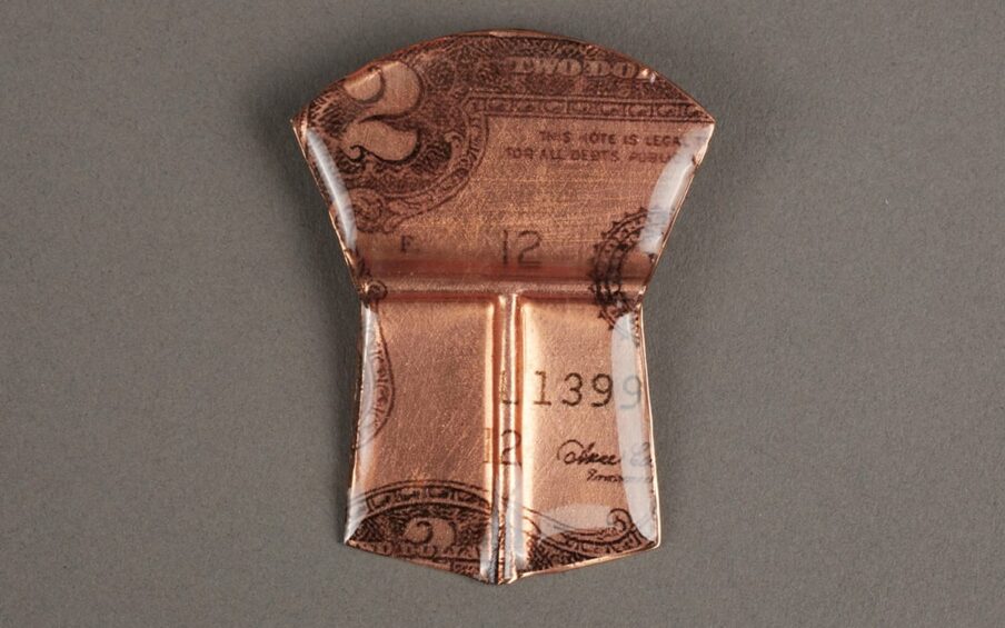 Image description: Two Dollars, Alison Bremner, resin and paper on copper, 2 x 1 ½ inches. A small shield-shaped object with a copper hue and details of a two dollar bill. The object is divided in two main parts: the upper and lower portions. The upper portion is shaped like a fan with a curved top that narrows at the sides and is divided from the lower part with a horizontal indentation in the glossy resin that covers the object. The fan contains the top left corner of a two dollar bill. A prominent number two is seen at left along with the elaborate scrollwork in the bill’s border. The words: “This Note Is Legal T…For All Debts Public” is seen at right. A capital letter “F.” and the number “12” are also seen on the plain lighter copper colored background along with part of a circular seal. Below the upper fan shape the shield is further divided in two with a vertical indentation. These two trapezoid shapes come to point at the center and show more of the bill including the numbers “1399” at right, a number “12” at center and a portion of the treasurer’s signature at lower right. The finish is glossy and smooth with beveled edges. the numbers “1399” at right, a number “12” at center and a portion of the treasurer’s signature at lower right. The finish is glossy and smooth with beveled edges.
