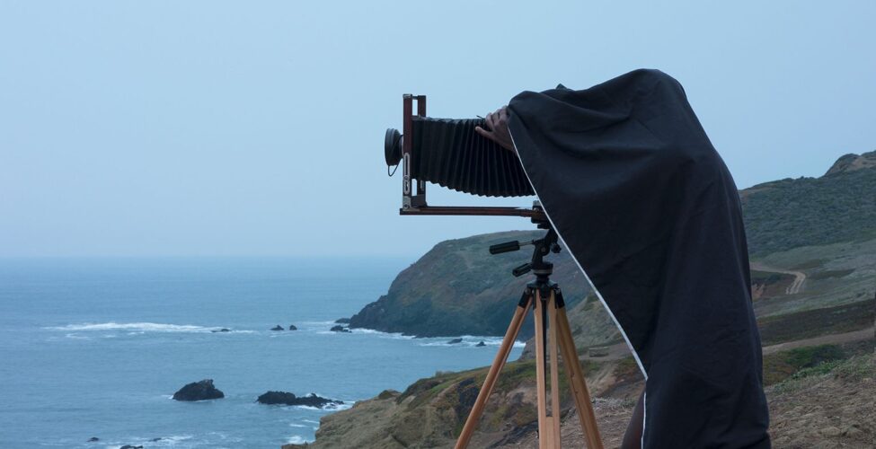 A vertical rectangular photograph of man using a camera on a tripod near an oceanside cliff.