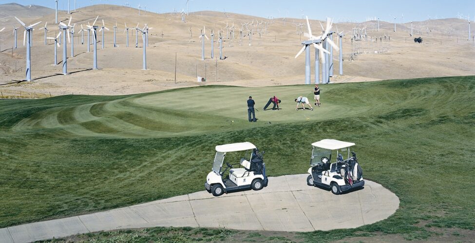 A landscape photograph of a golf course set amid bare, brown rolling hills with wind turbines.