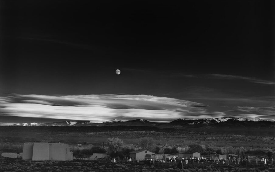 Image description: Moonrise, Hernandez, New Mexico, Ansel Adams, 1941, (negative) printed later, gelatin silver print. A black and white, landscape photograph showing a moon in a dark night sky over mountains, a church and graveyard. The photo is divided roughly in half with the top portion holding the dark, almost black sky. Towards the center of the work the sky lightens, and wispy clouds stretch across the photo. An almost full moon sits just left of center in the lighter sky above the light gray clouds. A snow-peaked mountain range sits under the clouds with scrubland below it. At far left a pueblo-style church with steep flat walls rises in the foreground topped with a small white cross. Low lying outbuildings and trees continue across to the right side of the photo where they meet with a cluster of crosses and headstones. Each cross is slightly different, and all seem to reflect the light and show up brightly against the darker background. Dark scrubby bushes continue to the bottom of the photograph.