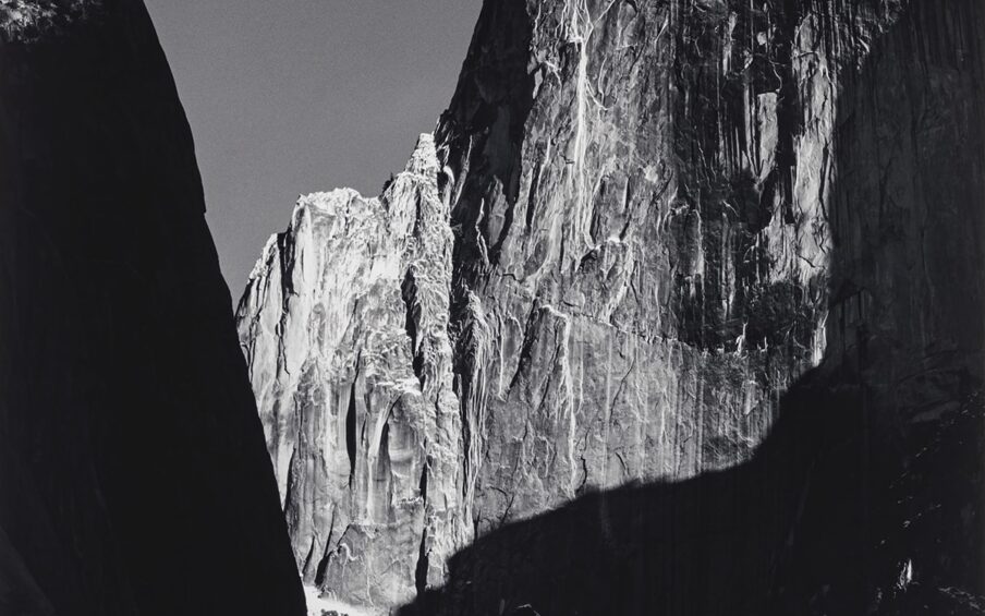 Image Description: Moon and Half Dome, Yosemite National Park. Ansel Adams. 1960. Gelatin silver print. Black and white photo of the sheer cliff wall of Half Dome in Yosemite under a waxing gibbous moon. The white moonlight reflects off the expansive Half Dome cliff edge highlighting the detailed texture in the rock appearing as vertical white lines. The jagged edges of the cliff are sharp and the top of the dome on the right appears smooth. A black edge of another cliff appears on the left side in the foreground. A large shadow in the shape of a backward L covers the right side of Half Dome extending from the right side of the photo into the middle. The rocks at the bottom of the two cliffs are slightly blurred and partially covered by shadows.