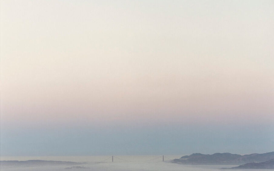 Image Description: Golden Gate Bridge 8.4.98 6:16am Richard Misrach. 1998. Photograph, chromogenic dye coupler print. Landscape photo showing the Golden Gate Bridge in the distance under the pastel colors of the sunrise. The bottom fourth of the photo shows peaceful bay waters covered with a light layer of fog. Dark peninsulas extend from either side of the bay just before the barely visible two pillars of the bridge. Calm water extends past the bridge and fades into the light blue sky. A thin layer of light purple extends beyond the blue followed by a light cream and blue sky.