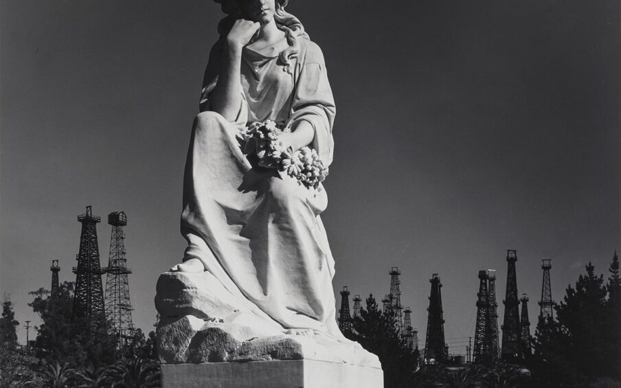 Image Description: Cemetery Statue and Oil Derricks, Long Beach, California. Ansel Adams. 1939. Photograph, gelatin silver print. Square black and white photo of a statue of a female with flowing wavy hair parted in the middle and gently falling onto her shoulders. Her eyes are closed and she has a peaceful, but solemn expression on her face. The figure is wearing a flowing robe-like garment with several ripples and folds. Her chin is gently resting on the folded fingers of her right hand and her elbow is resting on her knee. Her right foot and toes peek out from under the robe. She holds a wreath of daisy-like flowers in her left hand as it rests on her lowered left knee. The sculpture is on a cement pedestal that is partially visible with rounded corners decorated with filigrees. The figure seems illuminated while the evergreen trees, fifteen oil derricks, and sky in the background are much darker.