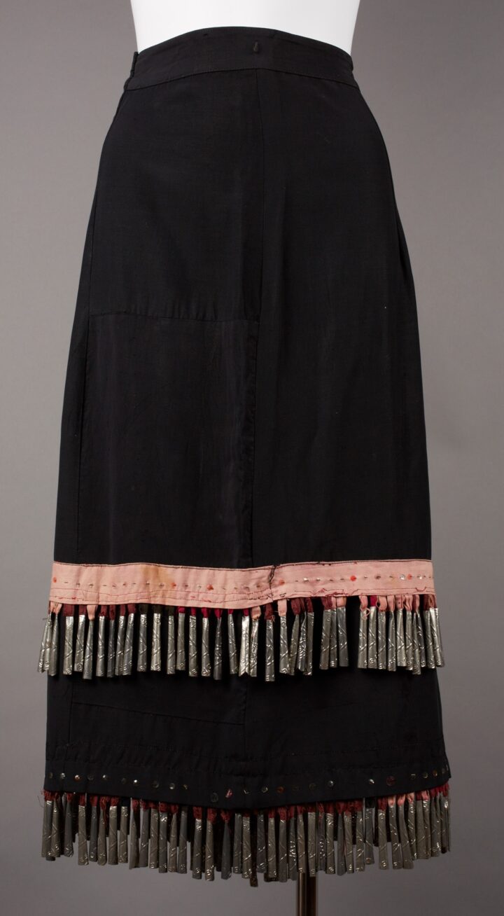 Ojibwa artist, Skirt, 27 x 31 ½ inches, cotton, plastic buttons, tin, and sequins. A long, black, slightly A- line shaped skirt decorated with two rows of metal jingles and a pale, peach stitched, fabric strip. The skirt has a flat waistband, fastens on the right side and falls in a slight flare to the hem. At lower calf level a faded peach colored strip of fabric bisects the skirt. It is embellished with a row of sequins in the middle and bordered by a line of black stitching at top and two lines of red stitching below. Jingles hang from under this strip from fabric loops in shades of red, brick, peach and black. The black skirt continues to the hem and finishes with another row of sequins on black with jingles on fabric loops extending from under the hem. The main body of the skirt is pieced in places, especially at the lower right where several rectangles of black fabric are stitched together to make up the garment. A center seam runs vertically down the skirt.