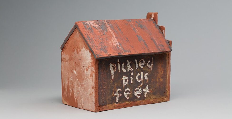 Image descriptions: Little’s Grocery (Shack #4), Willie Little, glazed ceramic, 9 ¾ x 7 ¾ x 10 ¾. 1. A three quarter view of a ceramic house with pentagon shaped ends, long sides and a sloped roof. The terracotta-colored roof has vertical ridges with deep gray and light beige portions creating a worn look. The broad side of the house is a dark brown with lighter shades of terracotta showing through. The words “pickled pigs feet” is painted on one broad side and “penny candy” on the other. The lettering is done in a lowercase, free hand style in a crackled, uneven, near white glaze with bits of terracotta showing through. The end of the house at right has a stairstep pattern rising from the gable, ending in a square shape at the peak. The short end of the house features lighter and darker terracotta colors that create texture and pattern.