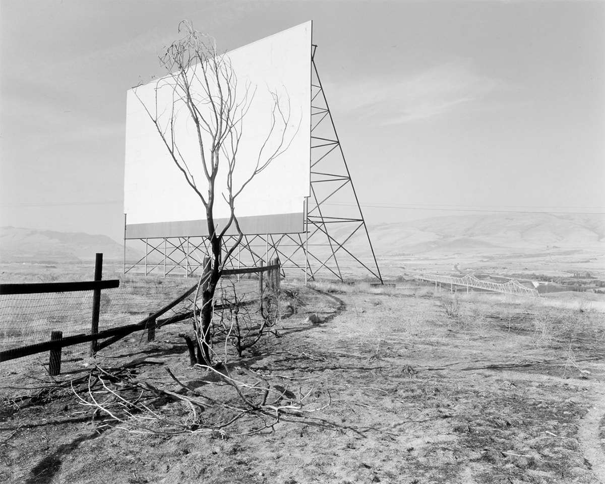 Image description: The Dalles, Oregon, Dan Powell, 1989, gelatin silver print, image: 12 11/16 x 15 7/8 inches; sheet: 16 x 19 7/8 inches. A black and white photograph featuring a drive-in movie screen and a bare tree amid an austere hilly, landscape. The movie screen occupies the upper left quadrant of the photo and shows the screen in a three-quarter view. A triangular structure supports the screen and its crossbar construction creates further triangular shapes. A small leafless tree stands near the screen and its darker branches cover most of the surface of the white screen. A dark wooden post and wire screen fence starts at center left and juts into the space behind the tree. The surrounding ground is strewn with bare branches and scrub grass. In the distance at right center and even with the bottom of the movie screen, the Dalles Bridge crosses a portion of the Columbia River. Low-slung, far-off hills bisect the photo in half. The top half of the photo shows a pale sky with light, wispy clouds.