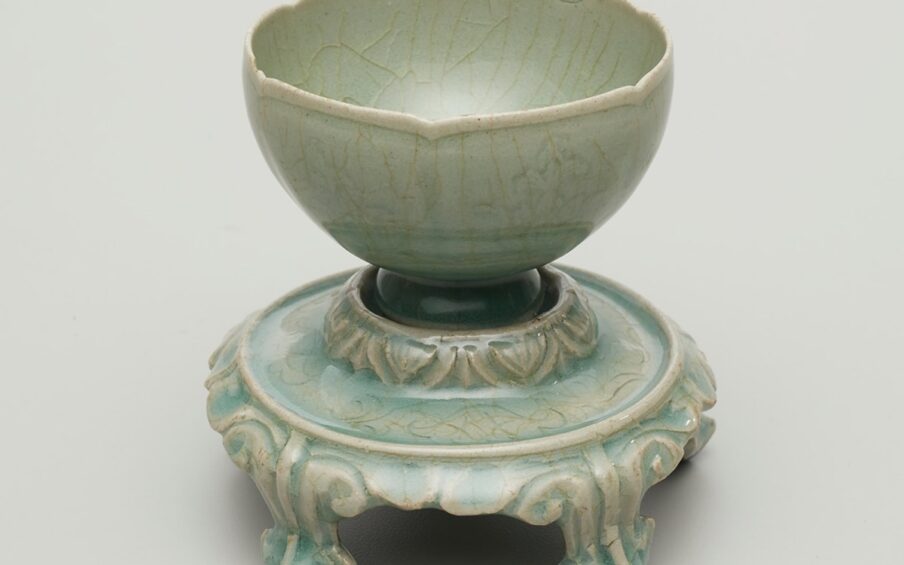 Image description: Foliated Cup and Stand with Floral Décor Korea, Jeollanam-do province, Gangjin kilns, Sadang-ri, light gray stoneware with incised and molded decoration under blue-green celadon glaze, 4 x 4 inches in diameter. A small round cup with a gently fluted rim and footed base sits on a round, decorated stand with five short legs. The cup and stand are a grayish-green with a crazed surface. The crazing creates a light brown, crackled pattern over the decorated surfaces of the work. The cup with its lip-rim mimicking a shallow flower petal edge, has molded floral pattern. The much narrower foot of the cup appears as a deeper green color and sits snugly in the raised stand. The stand’s upper tier is decorated with stylized V shapes before smoothly widening horizontally. A raised rim separates the upper tier from the five decorated legs of the stand. The legs end in a blunted point with two seeming to be too short to rest directly on the display surface.