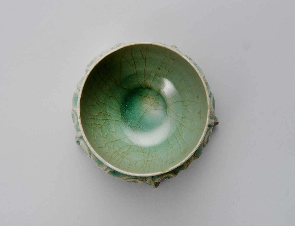 Image description: Foliated Cup and Stand with Floral Décor Korea, Jeollanam-do province, Gangjin kilns, Sadang-ri, light gray stoneware with incised and molded decoration under blue-green celadon glaze, 4 x 4 inches in diameter. A small round cup with a gently fluted rim and footed base sits on a round, decorated stand with five short legs. The cup and stand are a grayish-green with a crazed surface. The crazing creates a light brown, crackled pattern over the decorated surfaces of the work. The cup with its lip-rim mimicking a shallow flower petal edge, has molded floral pattern. The much narrower foot of the cup appears as a deeper green color and sits snugly in the raised stand. The stand’s upper tier is decorated with stylized V shapes before smoothly widening horizontally. A raised rim separates the upper tier from the five decorated legs of the stand. The legs end in a blunted point with two seeming to be too short to rest directly on the display surface.