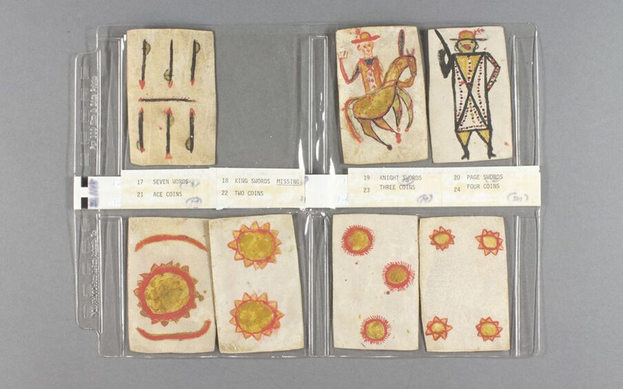 Image description: Playing Cards, Apache artist, colored ink on paper, 35 cards, each 3 ½ x 2 ¼ inches. Seven handmade playing cards decorated in bright reds and yellows displayed and labeled on a clear plastic holder. The top row of cards shows “Seven Swords” with three vertical swords over another three bisected by a horizontal sword. Next, a space is left for a missing card, “King Swords”. “Knight Swords” is next and shows a figure wearing a hat and yellow and red uniform riding a brown horse. “Page Swords” is last in the row; The figure wears a plumed hat, and its body is depicted as a long rectangle with a black X across it. Red and black dots decorate the body with smaller triangles making up the figure’s legs. The bottom row of cards starts with “Ace Coins” shown at a large yellow circle in center ringed with red and edged in small yellow triangles outlined in red. Two red semi circles bracket the yellow coin on top and bottom. The next card is “Two Coin”, two stacked yellow coins, rimmed with triangles and outlined in red. “Three Coins” shows the same style of coins positioned at 11, 3 and 6 o’clock. The last card in the bottom row is “Four Coins” and it uses the same yellow and red coin show one in each corner of the card. The cards rest on a clear plastic holder and paper labels separate the top and bottom rows.