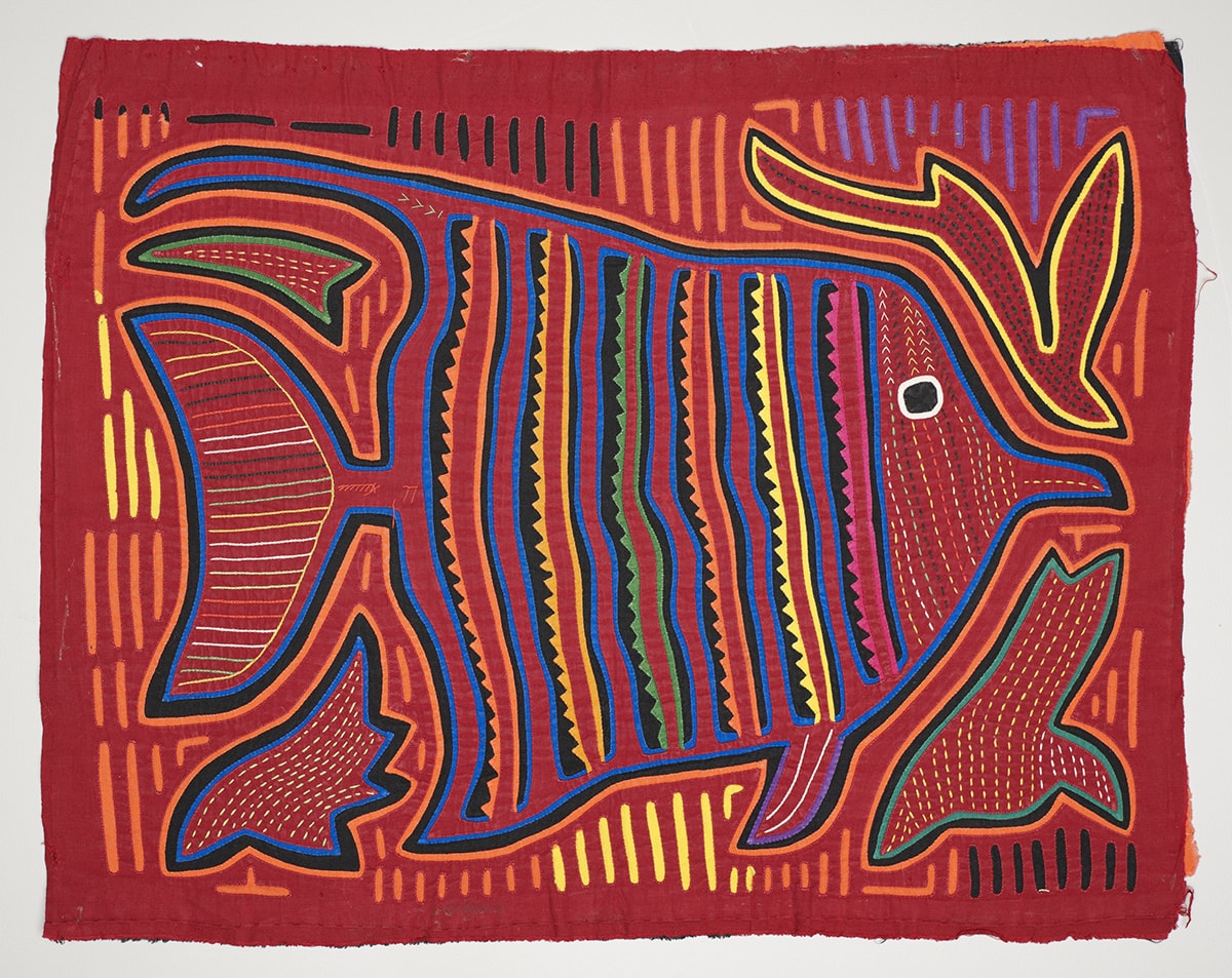 Image Description: Fish Mola. Kuna artist. Cotton. 13 ¾ in x 17 ½ in. Bright red fabric with a fish embroidered on it. The fish is outlined in a thick black line with a bright blue line against it on the inside and a bright orange line on the outside. The fish is triangular shaped and facing the right. It has a large flat tail and small fins in front. Its mouth is small and beak shaped. It has a large black eye with a white outline. Its face is a series of lines of small dots that are yellow and red. Behind the eye, solid colorful lines: blue, black, pink, red, pink, scalloped black, blue, red, blue, black, yellow, red, yellow, scalloped black, blue, red, blue, black, orange, red, orange, scalloped black, blue, red, blue, black, green, red, green, scalloped black, blue, red, blue, black, yellow, red, yellow, scalloped black, blue, red, blue, black, orange, red, orange, scalloped black, blue. The tail has several horizontal lines of varying colors (green, yellow, white, red, orange). There are also lines of various colors between the outline of the fish and the edge of the fabric (black, yellow, orange, and purple). There are four shapes at the corners of the fish. The top left is a triangular shape with a green and black outline and yellow dots. The top right is a shape that resembles a bird with a long tail diving downward. It is outlined in yellow and black and has dark spots. The bottom right also resembles a bird diving and is outlined in green and black with yellow spots inside. The bottom left is flame shaped with blue and black outlines and yellow dots inside.