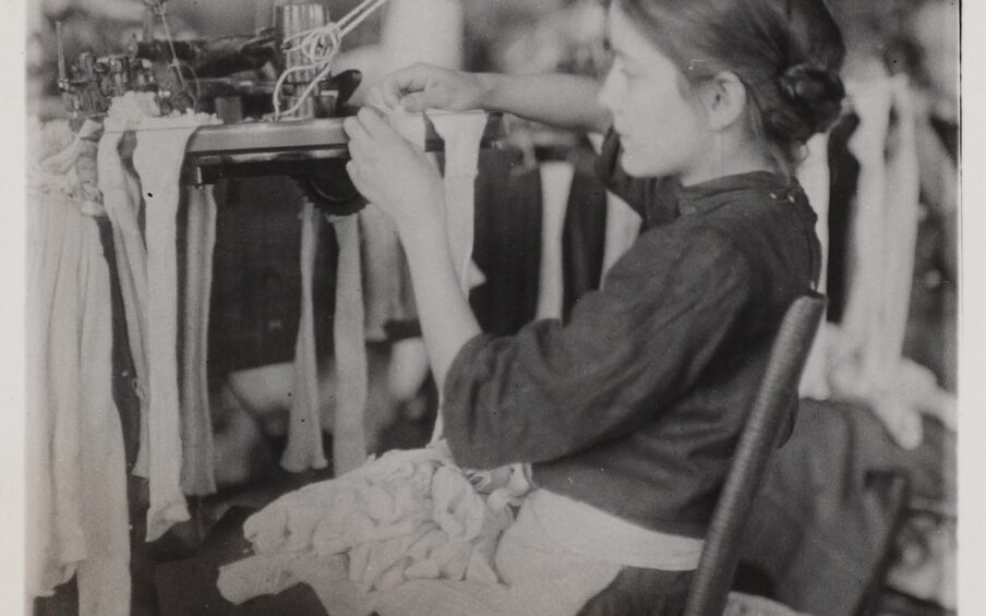 Image description: Lewis Hine, Making Stockings, gelatin silver print, 4 1/2 x 31/2 in. Vertical rectangular black-and-white photo of a young girl in profile sitting on a wood chair slightly tilted back that faces left, her hands and quiet concentration focused on a factory machine with stockings draped from it. She wears a dark dress, light apron, her hair appears to be in a low bun, and her lap is full of stockings which spill over to the floor and create a heap to her right. The background is out of focus and suggests disorderly conditions.