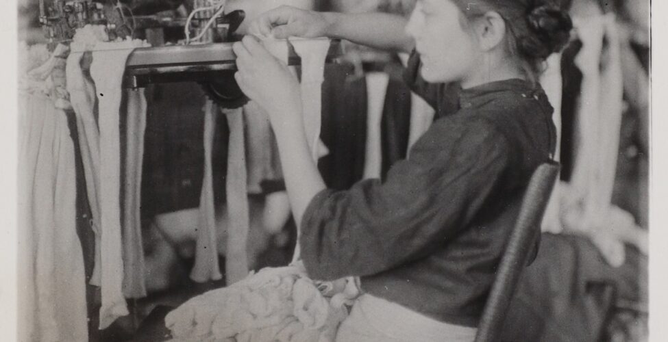 Image description: Lewis Hine, Making Stockings, gelatin silver print, 4 1/2 x 31/2 in. Vertical rectangular black-and-white photo of a young girl in profile sitting on a wood chair slightly tilted back that faces left, her hands and quiet concentration focused on a factory machine with stockings draped from it. She wears a dark dress, light apron, her hair appears to be in a low bun, and her lap is full of stockings which spill over to the floor and create a heap to her right. The background is out of focus and suggests disorderly conditions.