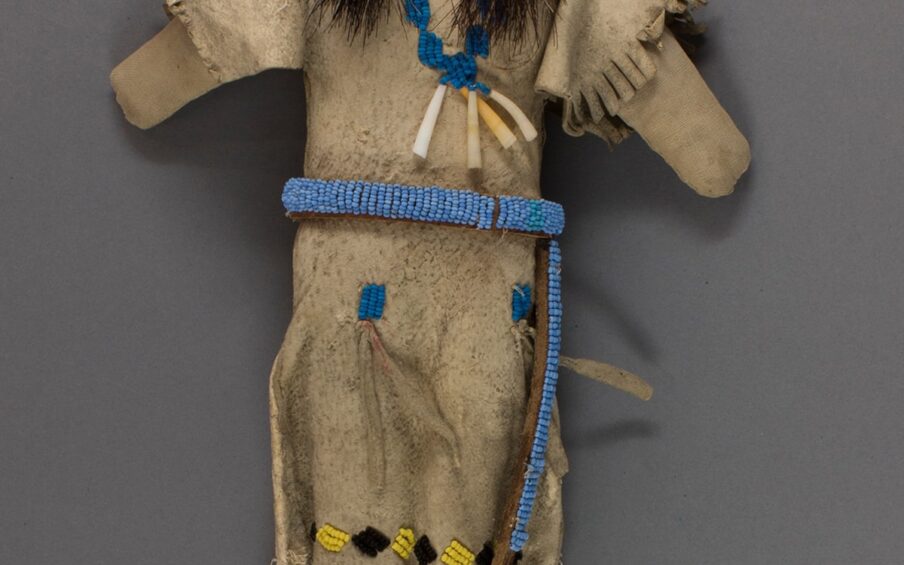 Image description: Doll, Nez Perce artist, hide, beads, cloth and hair, 12 x 7 3/4 x 1 1/8 inches. A cloth doll clothed in a beaded hide dress with dark brown hair in pigtails. The doll’s creamy tan face shows wear and has beaded features. Single black beads sandwiched between two white beads compose the eyes, two black beads make up the nostrils and a longer line of red beads topping a shorter row make the mouth. Brown hair is gathered in two pigtails with yellow beads on either side of the head. The doll’s arms stand straight out from the body and have rounded ends representing hands. The doll wears a long hide dress in the same creamy tan as the body that has fringed cuffs and hem. A bright blue beaded necklace with four long peach and white shells is stitched around the neck. A sky-blue beaded belt with long ties wraps around the waist. Two bright blue beaded patches at the hips have hide tassels. Alternating yellow and black beaded diamonds appear just above the fringed hem. The doll wears hide leggings embellished with sky-blue beads and moccasins beaded in yellow and bright blue. The doll appears worn and much loved.