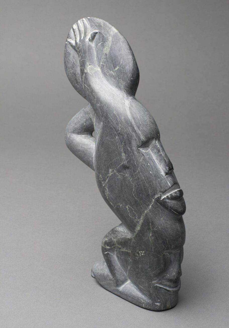 Image Description: Spirits. Silas Aittauq. 1986. Serpentine carving 9 in x 4 1/2 in x 1 3/4 in. Sleek gray carving of a spirit with multiple faces. From the angle of the photograph - the body is facing to the right, but the head is turned to face the camera. The face is round with almond shaped eyes and a thin nose carved into it. A slightly opened mouth lies behind a raised strip under the nose. It has bangs that are parted in the middle and angle to each side of the head. The body is leaning forward, right arm bent at the elbow with the hand holding the chin of the turned face. The legs are bent at the knees and there are rounded feet below. The back and space behind the legs has the profile of two additional faces. Long thin noses and plump lips are visible. There are blemishes in the material and scratches across the sculpture. A prominent light colored scratch runs from the right eye, over the lips and chin, over the finger, and down to the armpit.