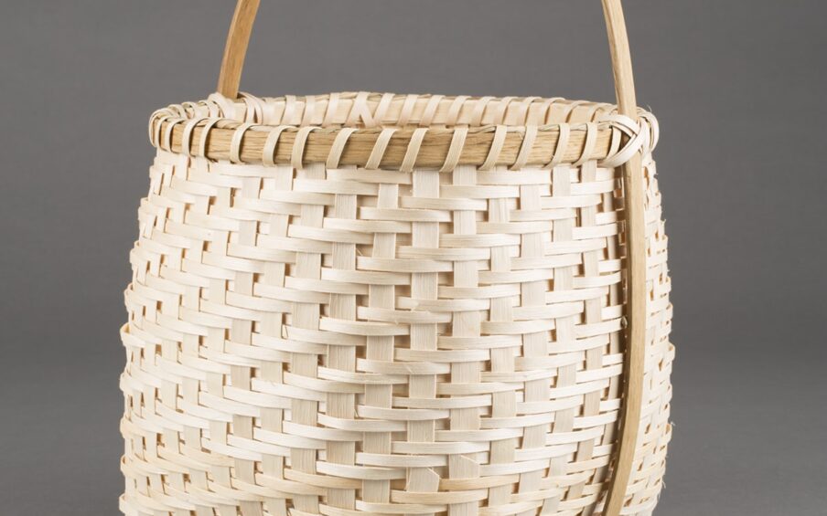 Image Description: Honor the Harvest (Corn Washing Basket). Nan MacDonald. 2014. White ash, sweetgrass, and oak basket. 18 in x 12 in diameter. This tall basket is square on the bottom with pointed bases on the four sides and has a rounded top with a smooth wooden handle that runs over the top diagonally and down both sides, connecting on the bottom of the basket. The intricate woven pattern has larger strips of light wood evenly spaced vertically with two thin horizontal bands connecting them. The smooth wooden lip at the top has a darker grain and then thin strips which are angled diagonally and evenly spaced. They cross the handle against the lip in an x shape.