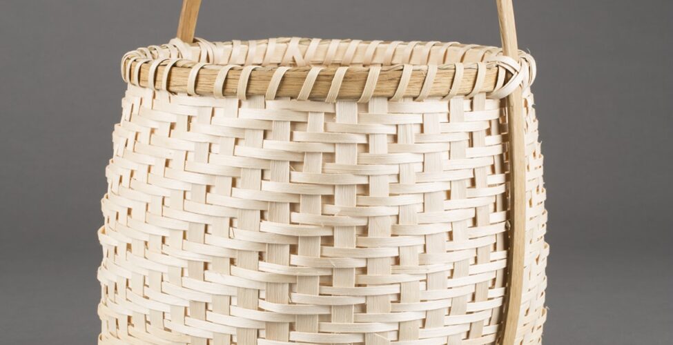 Image Description: Honor the Harvest (Corn Washing Basket). Nan MacDonald. 2014. White ash, sweetgrass, and oak basket. 18 in x 12 in diameter. This tall basket is square on the bottom with pointed bases on the four sides and has a rounded top with a smooth wooden handle that runs over the top diagonally and down both sides, connecting on the bottom of the basket. The intricate woven pattern has larger strips of light wood evenly spaced vertically with two thin horizontal bands connecting them. The smooth wooden lip at the top has a darker grain and then thin strips which are angled diagonally and evenly spaced. They cross the handle against the lip in an x shape.