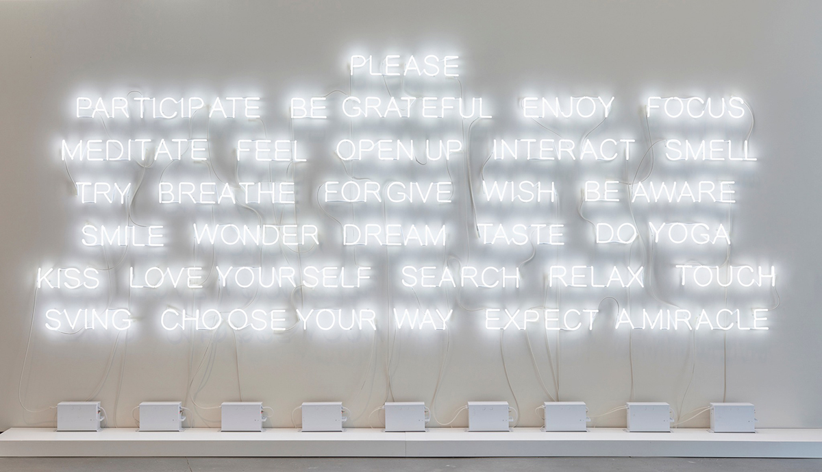 Image description: Please Participate. Jeppe Hein. 2015. Neon tubes and transformers. 88 inches x 210 inches. Words in white neon mounted on a white gallery wall in seven rows. The lettering is simple and approachable in all capitals. The words are: Row 1: Please. Row 2: Participate, Be, Grateful, Enjoy, Focus. Row 3: Meditate, Feel, Open, Up, Interact, Smell. Row 4: Try, Breathe, Forgive, Wish, Be, Aware. Row 5: Smile, Wonder, Dream, Taste, Do, Yoga. Row 6: Kiss, Love, Yourself, Search, Relax, Touch. Row 7: Sving, Choose, Your, Way, Expect, A, Miracle. Below nine, white, rectangular transformers sit on a shelf below the words with electrical cords connecting each one to several words. The upper left corner of the image shows an opening in the wall and light from the gallery above.