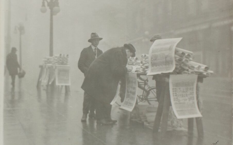 Image description: The New Year’s Edition (of the Oregonian), William H Walker, gelatin silver print, image 7 x 8 3/4 inches, sheet 7 1/16 x 9 ¼ inches. This black and white photograph depicts a newspaper vendor and customers at an outdoor newsstand on the sidewalk of a rainy street. At center right, a makeshift table holds mounds of large rolls of newspaper broadsheets. Three oversized broadsheets hang from the table and are half the length of the figure bent over the table. The figure, standing at the photo’s center, wears long dark overcoat and trousers with a hat. Behind them and to the left stands another figure, again in dark clothing and hat. The newsstand sits on the angled sidewalk which runs from the lower right to the upper left of the photo. Further down the sidewalk at far left is a streetlamp and another newsstand table heaped with rolled papers. In the distance, at far left another dark clad figure approaches. The background appears misty and the sidewalk shines with the fallen rain. At right, the hazy outline of buildings across the street are just discernible.