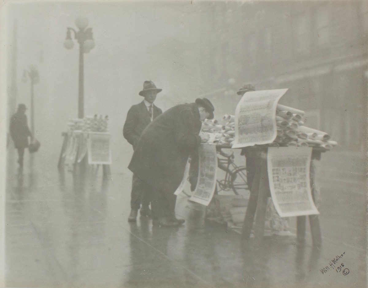 Image description: The New Year’s Edition (of the Oregonian), William H Walker, gelatin silver print, image 7 x 8 3/4 inches, sheet 7 1/16 x 9 ¼ inches. This black and white photograph depicts a newspaper vendor and customers at an outdoor newsstand on the sidewalk of a rainy street. At center right, a makeshift table holds mounds of large rolls of newspaper broadsheets. Three oversized broadsheets hang from the table and are half the length of the figure bent over the table. The figure, standing at the photo’s center, wears long dark overcoat and trousers with a hat. Behind them and to the left stands another figure, again in dark clothing and hat. The newsstand sits on the angled sidewalk which runs from the lower right to the upper left of the photo. Further down the sidewalk at far left is a streetlamp and another newsstand table heaped with rolled papers. In the distance, at far left another dark clad figure approaches. The background appears misty and the sidewalk shines with the fallen rain. At right, the hazy outline of buildings across the street are just discernible.