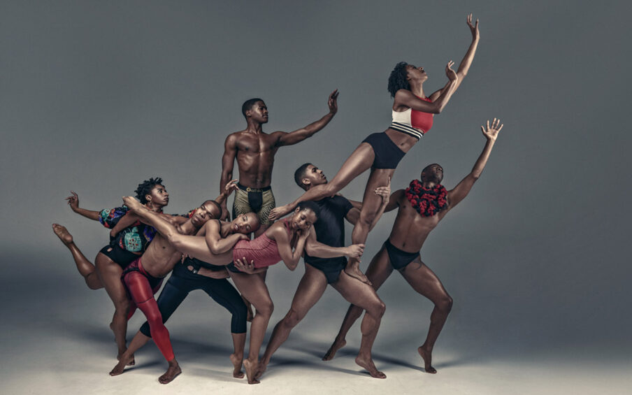 Image Description: Photo of eight Black dancers posing in a multi-level human sculpture on a gray stage in bright white light. Four dancers form a foundation. One in the center with black calf-long leggings and a black tank top stands with legs apart, toes pointed. To the right another dancer wears a red tank top and black leotard. They stand on their left toe with their right leg extended behind. The dancers behind embrace the leg and rest their heads on it. To the left of the central dancer with legs apart is a dancer with bright red leggings. Their left foot is extended behind and right foot forwarded and angled to the right. They lay their head on the center dancer’s shoulder. To the left is a dancer wearing a black leotard and a brightly colored shirt with puffy sleeves. It has multiple rounded rectangles in teal, blue, and red, with white and red accents. The dancer stands on their right leg, left leg bent with toes extended behind their back, with their arms outstretched and fingers gently curved. Behind the line of dancers, a single dancer wearing light gold shorts with black dots stands on their toes, facing the audience with left arm extended, palm out, fingers raised upward, but gently bent. At the right end of the line of dancers is a dancer wearing a black leotard with short sleeves. They stand facing the right with legs apart, toes pointed. They hold another dancer on their left leg. This dancer’s right leg extends over the base dancer’s shoulder as that dancer holds the left thigh and ankle. The dancer looks upward, wearing black briefs and a red sports bra with white sides and white and black striped base, leans to the right, left arm extended, right arm bent at the elbow with palms facing out and fingers gently bent. Behind, a dancer on the ground wearing a black speedo leotard and a large puffy black and red scarf holds the shoulder of the dancer in the full black leotard with their right arm and extends their left arm upward, palm facing forward, fingers extended.