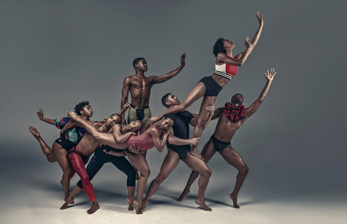Image Description: Photo of eight Black dancers posing in a multi-level human sculpture on a gray stage in bright white light. Four dancers form a foundation. One in the center with black calf-long leggings and a black tank top stands with legs apart, toes pointed. To the right another dancer wears a red tank top and black leotard. They stand on their left toe with their right leg extended behind. The dancers behind embrace the leg and rest their heads on it. To the left of the central dancer with legs apart is a dancer with bright red leggings. Their left foot is extended behind and right foot forwarded and angled to the right. They lay their head on the center dancer’s shoulder. To the left is a dancer wearing a black leotard and a brightly colored shirt with puffy sleeves. It has multiple rounded rectangles in teal, blue, and red, with white and red accents. The dancer stands on their right leg, left leg bent with toes extended behind their back, with their arms outstretched and fingers gently curved. Behind the line of dancers, a single dancer wearing light gold shorts with black dots stands on their toes, facing the audience with left arm extended, palm out, fingers raised upward, but gently bent. At the right end of the line of dancers is a dancer wearing a black leotard with short sleeves. They stand facing the right with legs apart, toes pointed. They hold another dancer on their left leg. This dancer’s right leg extends over the base dancer’s shoulder as that dancer holds the left thigh and ankle. The dancer looks upward, wearing black briefs and a red sports bra with white sides and white and black striped base, leans to the right, left arm extended, right arm bent at the elbow with palms facing out and fingers gently bent. Behind, a dancer on the ground wearing a black speedo leotard and a large puffy black and red scarf holds the shoulder of the dancer in the full black leotard with their right arm and extends their left arm upward, palm facing forward, fingers extended.