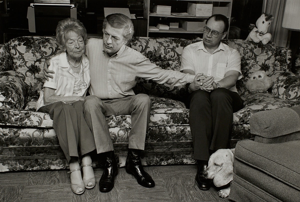 Sage Sohier, Gordon and Jim with Gordon’s Mother Margot, 1987, pigment print, 15 9/16 x 23 in. horizontal b&w photo with hot flash depicting 3 figures seated on a floral couch, the central male with his right arm around the elderly woman and left arm reaching out to male seated further away who is grasping it in both hands.