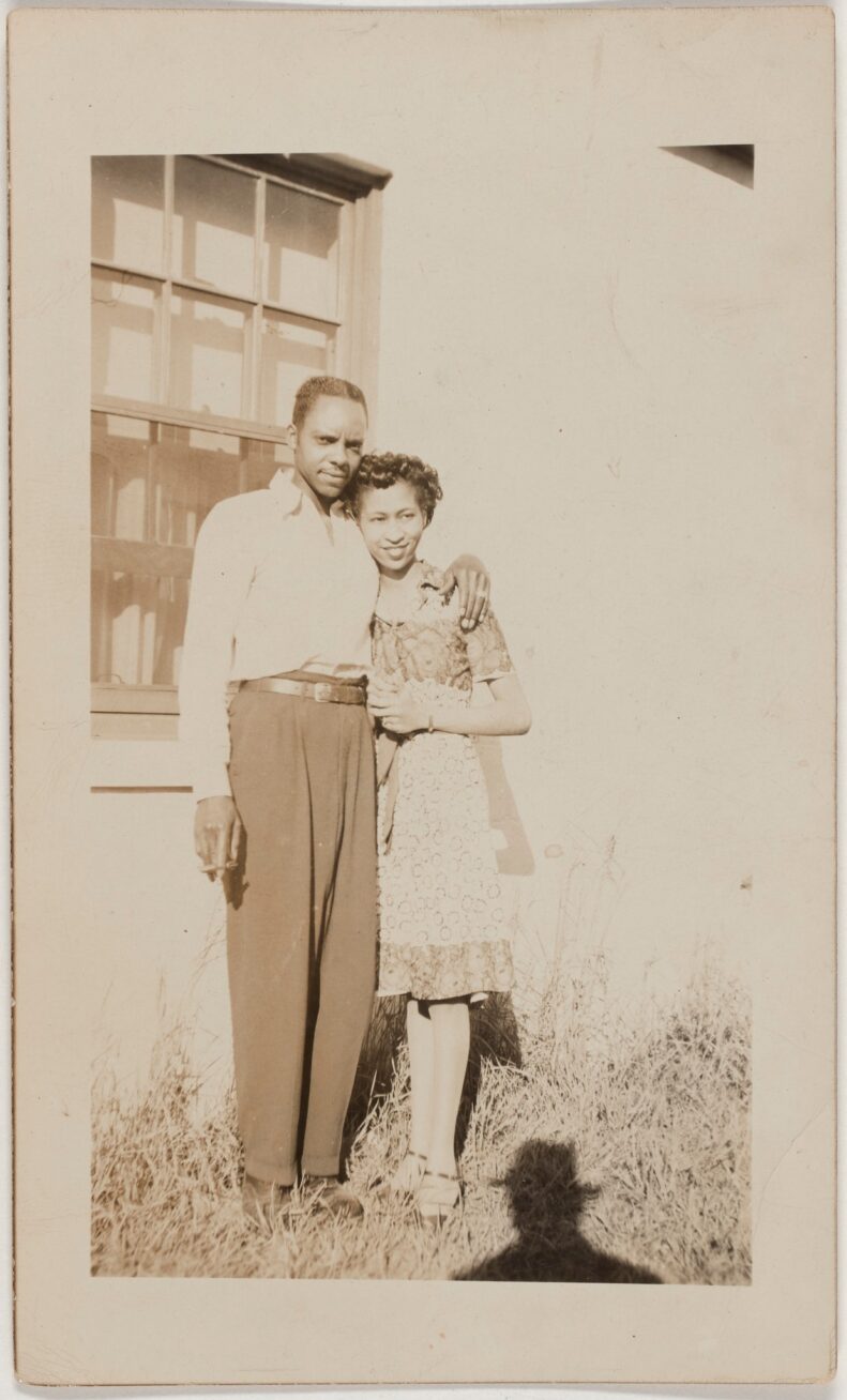 Unknown photographer, Snapshot of Couple Standing in Front of Window, 1960/65, gelatin silver print, 4 ¾ x 2 ¾ in, sepia-toned outdoor vernacular photo of a couple wearing vintage clothes in front of a window. Male figure wears white shirt and dark slacks and holds a cigarette in his right hand, left hand around the shoulder of a woman in a patterned dress. Photographer’s shadow is visible in bottom of frame.