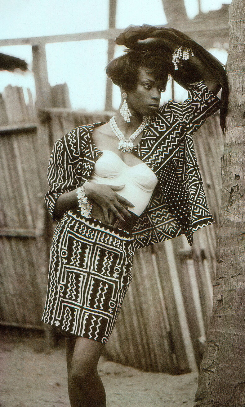 Black and white photograph of a black model posing against a tree, wearing a white bustier and a matching skirt and jacket and pearl jewelry