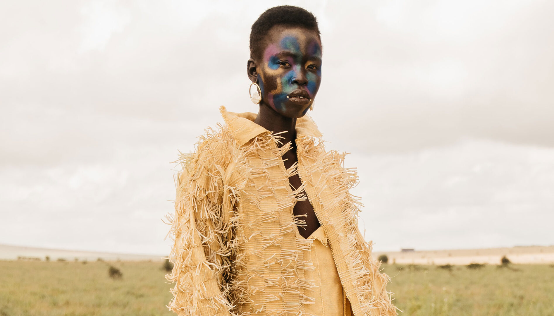 Photo of a black model with iridescent face paint and a yellow fringe jacket standing against a cloudy sky and grassy landscape