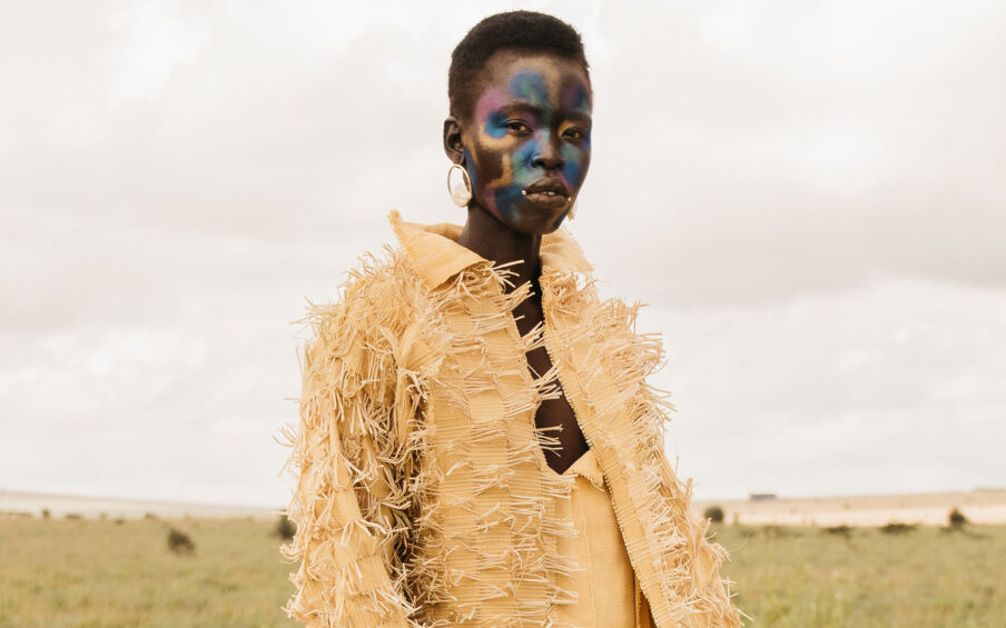 Photo of a black model with iridescent face paint and a yellow fringe jacket standing against a cloudy sky and grassy landscape