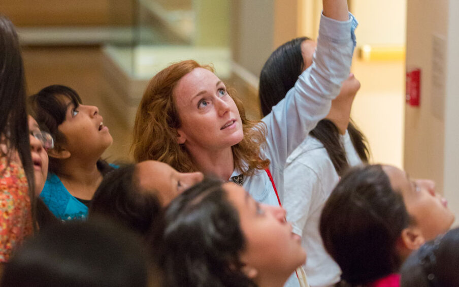 Photo of a woman with long red hair pointing upwards towards an artwork, surrounded by a group of students
