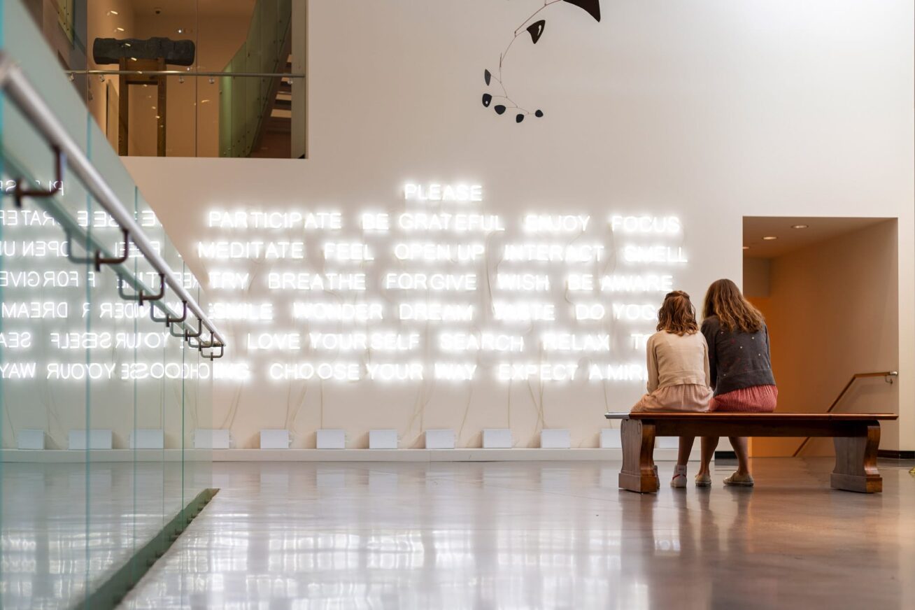 Two people sitting on a bench viewing an installation of neon text on a wall
