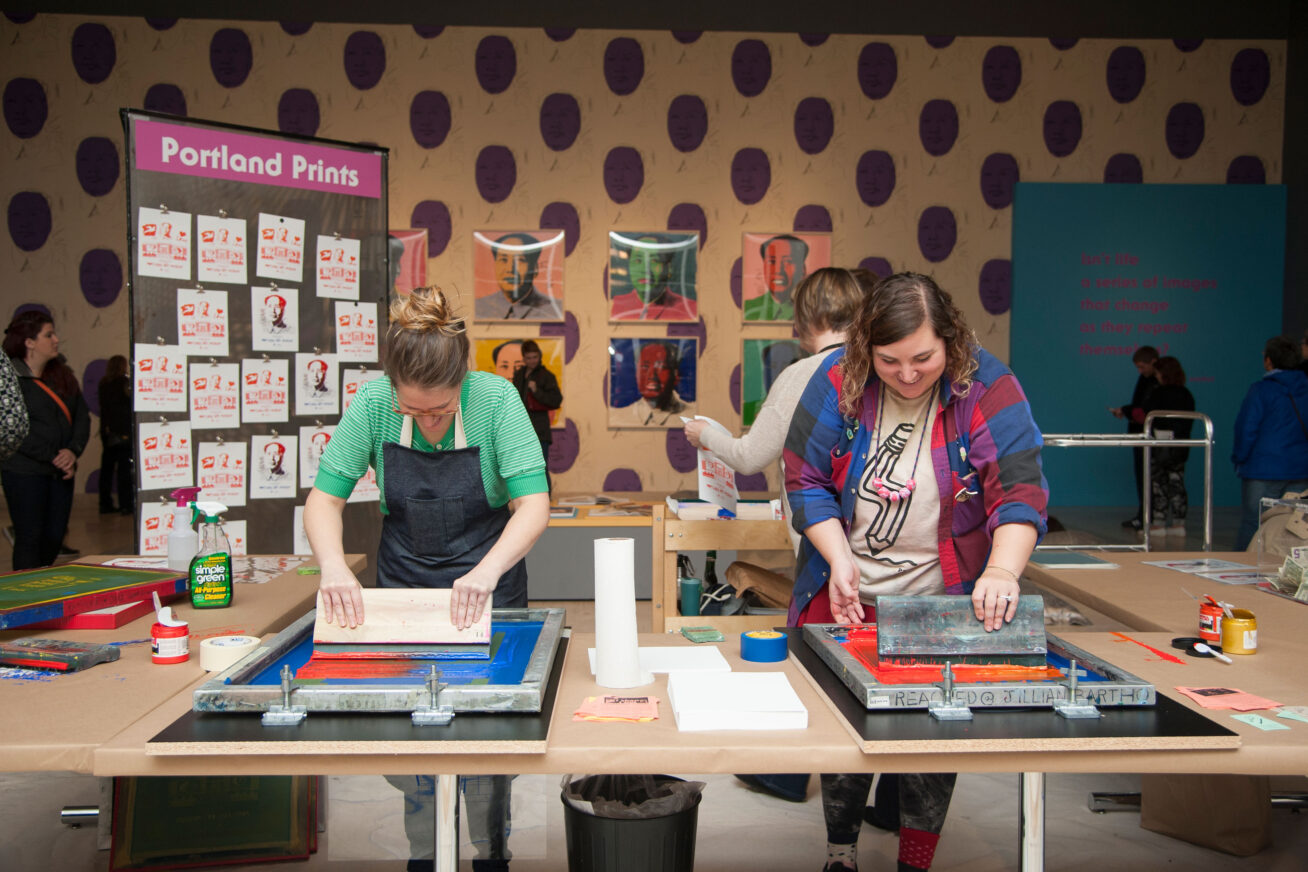 Color photo of two smiling women, one wearing an apron, making screenprints. In the backgrounds, images of Andy Warhol's Mao screenprints on a purple polka dot wall.