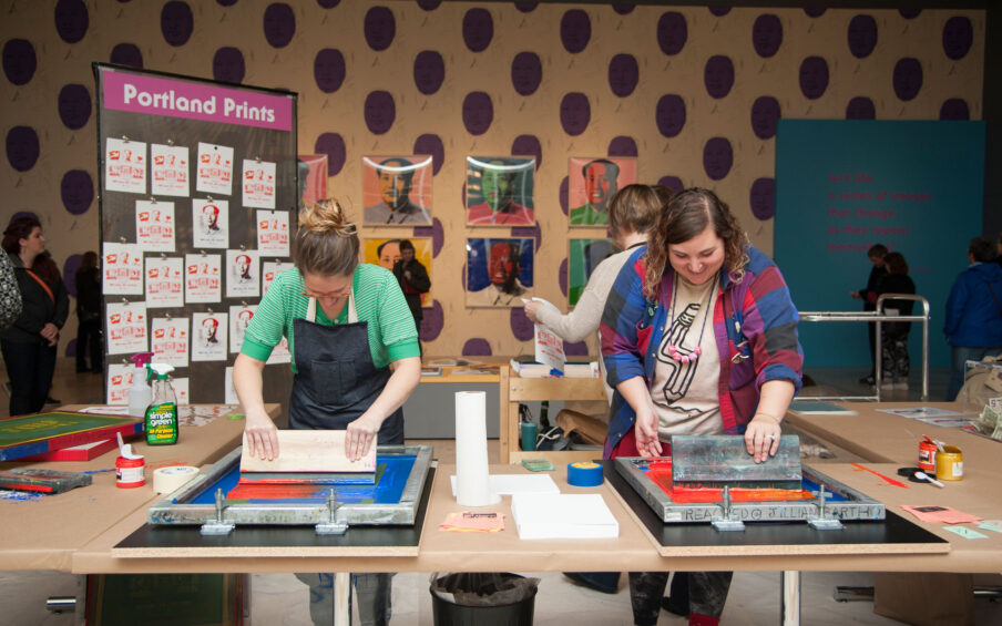 Color photo of two smiling women, one wearing an apron, making screenprints. In the backgrounds, images of Andy Warhol's Mao screenprints on a purple polka dot wall.