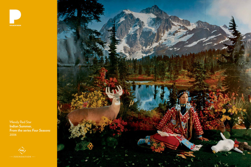 Poster with image of Wendy Red Star's "Indian Summer," from the series "Four Seasons." Image of a Native American woman in a traditional dress with beaded bag and accessories in front of an image of a nature scene with mountains, a cardboard cutout of a deer, and a fake cow skull in the foreground.