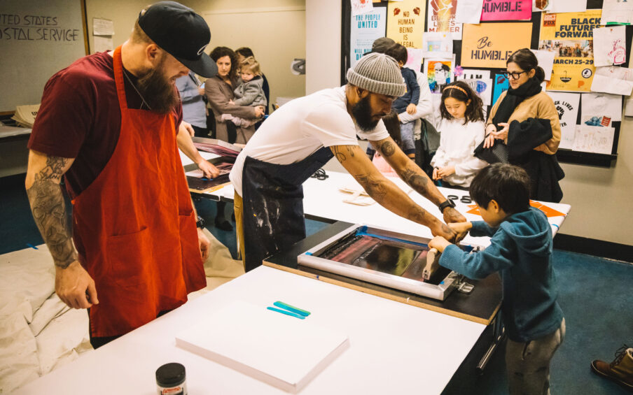 Photograph of a man with a beanie cap and a black apron on helping a young boy make a screenprint. Parents and children and another man in a red apron and black baseball hat look on in the background.