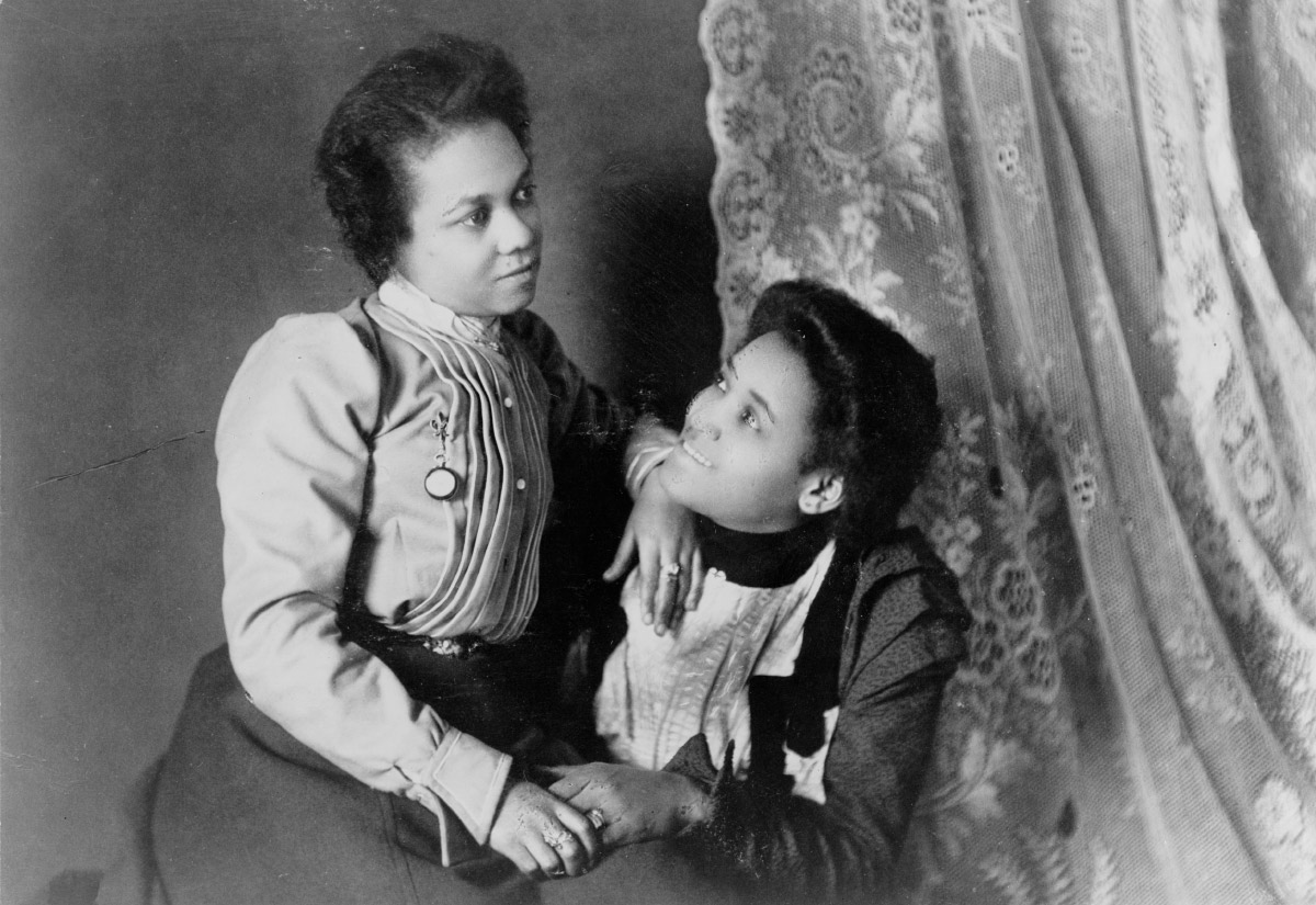 Old photo of one Black woman sitting on another Black woman's lap who is smiling up at her, both wearing late-nineteenth century clothing
