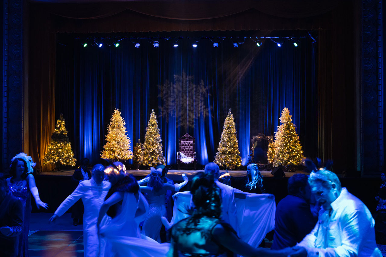 Photo of a crowd of people dancing, wearing white clothing. In the back, a dark stage, with lit up Christmas trees and a throne in the middle.