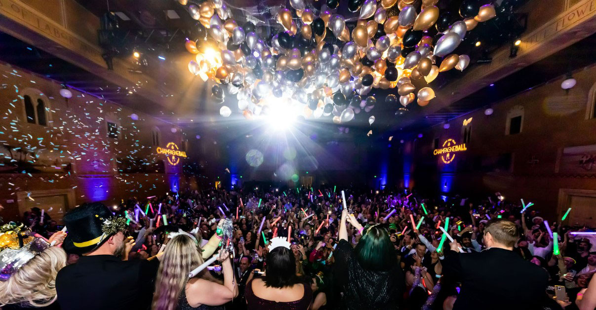Color photograph of a celebration in the Sunken Ballroom of the Museum. Balloons dropping from the ceiling and a big crowd of people below with glow sticks and party hats on.