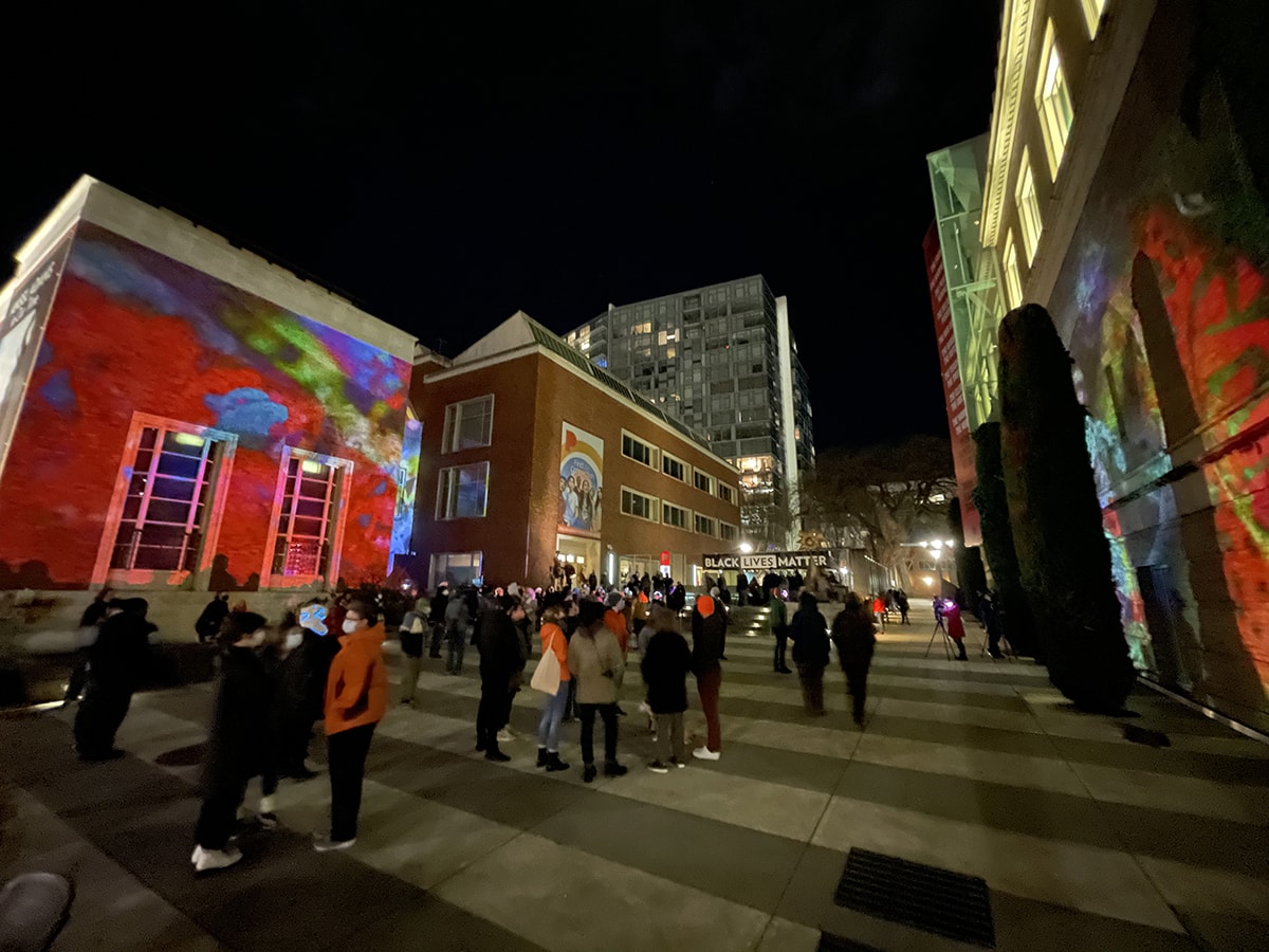 The exterior courtyard of the Portland Art Museum at night with video being projected on the building and people gathered in the courtyard